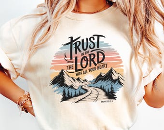May include: A white t-shirt with a graphic design featuring a mountain landscape with a road winding through it. The text "TRUST IN THE LORD WITH ALL YOUR HEART" is written in a stylized font with a sunset background. The text "PROVERBS 3:5" is written below the graphic.