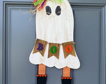 May include: A white wooden ghost wall hanging featuring black oval eyes and a brown banner with the letters B, O, and O in purple, green, and orange. The ghost wears orange and white polka dot legs with black boots and a colorful fabric bow.