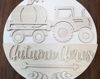 May include: A circular wooden sign featuring a light-colored finish. The top half displays a laser-cut tractor pulling a wagon with a pumpkin. The bottom half features the text Autumn Acres in script font, an arrow, and two leaf sprigs.