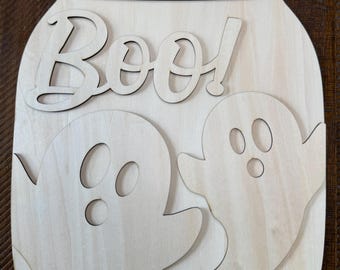 May include: A light-colored wooden cutout shaped like a mason jar featuring the word Boo! in a script font at the top. Two ghost figures with cut-out eyes and mouths are layered onto the lower portion of the jar. The top rim has two holes.
