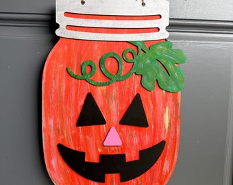 May include: A hanging wall decoration shaped like a mason jar painted in orange with a distressed finish. It features a jack-o'-lantern face with black triangular eyes, a pink triangular nose, a black jagged mouth, and a green vine with a leaf.