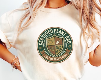 May include: Cream-colored sweatshirt with a circular graphic. The graphic reads "CERTIFIED PLANT DAD" in green and white, with a shield design featuring gardening tools and plants. Below the graphic, it says "I WET MY PLANTS DAILY".