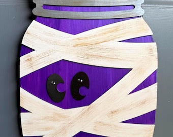 May include: A wooden wall hanging shaped like a mason jar painted purple with a silver lid. The jar is wrapped in off-white, distressed-look bandages and features two black, crescent-shaped eyes with small white dots, suspended by a brown twine string.