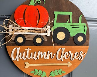 May include: A circular wooden door sign featuring a dark brown background with a bright orange pumpkin in a white wagon, a green tractor, and the text Autumn Acres in white script. Below the text is a horizontal arrow and green leaf accents.