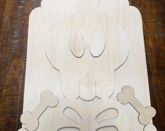 May include: A light-colored wooden craft cutout featuring a skeleton design. The piece includes a rectangular backing with a rounded top and a hole for hanging, with separate wooden skull, rib cage, and bone-shaped pieces arranged on top.