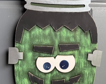 May include: A decorative wooden wall hanging shaped like a mason jar, painted green with a black jagged hairline, black eyebrows, a black mouth with teeth, and two large white eyes with dark blue pupils. It features silver side bolts and a lid.