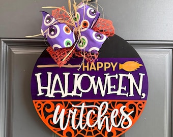 May include: A round wooden door sign featuring a purple and orange color scheme. The text reads HAPPY HALLOWEEN, witches in white lettering. A broomstick graphic is included. The top is decorated with a purple ribbon with eye patterns.