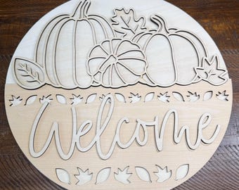 May include: A circular wooden sign featuring a layered design with three pumpkins and several leaves at the top. The word Welcome is written in a cursive script across the center, with additional leaf cutouts positioned above and below the text.