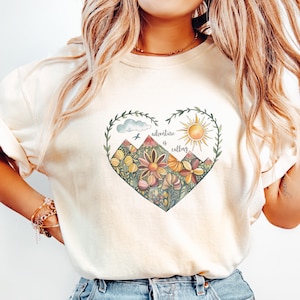 Puede incluir: Una camiseta blanca con un diseño en forma de corazón que presenta montañas, un sol, una nube y flores. El texto "adventure is calling" está escrito dentro del corazón.