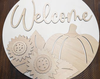 May include: A circular wooden sign featuring a light-colored background with the word Welcome in cursive script. The sign is decorated with layered wooden cutouts of a pumpkin and two sunflowers with leaves, all in a natural light wood finish.