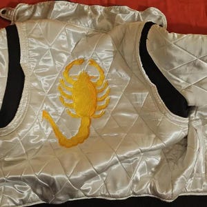 Puede incluir: Una chaqueta de satén blanco acolchado con un gran escorpión amarillo bordado en la espalda. La chaqueta tiene ribetes negros acanalados en el cuello y las aberturas de las mangas, con un patrón de costuras en rombos sobre la tela brillante.
