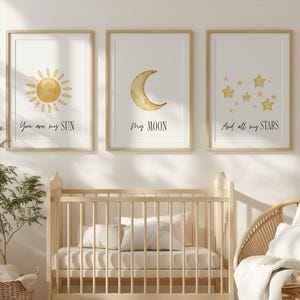 May include: Three framed art prints with watercolour illustrations of a sun, moon, and stars, with the phrases "You are my SUN", "My MOON", and "And all my STARS". Below, a wooden cot, a woven chair, and a round rug.