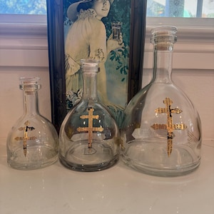 May include: Three clear glass bottles with cork stoppers and gold cross designs. The bottles vary in size, with a vintage Coca-Cola advertisement in the background. The bottles are likely for decorative purposes or storing liquids.