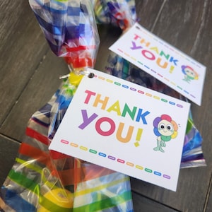 Puede incluir: Dos etiquetas de agradecimiento coloridas con un diseño de arcoíris y el texto "¡Gracias!" en una fuente de arcoíris. Un personaje de flor de dibujos animados está en el lado derecho de cada etiqueta.