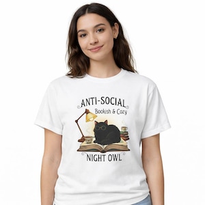 Op de afbeelding: Wit T-shirt met een zwarte kat met een bril, zittend op een boek, met een lamp en koffiekopjes. De tekst op het shirt luidt "ANTI-SOCIAL Bookish & Cozy NIGHT OWL". Een gezellig ontwerp voor boekenliefhebbers.