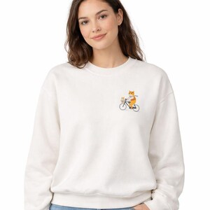 May include: A cream-colored sweatshirt featuring an embroidered design of a fox riding a bicycle with a smaller fox in the front basket. The fox is wearing an orange outfit and holding a baguette. The bicycle is black.