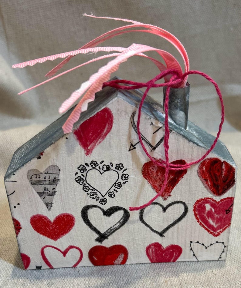 May include: A small wooden house shaped ornament with a gray painted roof and a chimney. The house is decorated with a red and black heart pattern and a pink ribbon tied around the chimney.