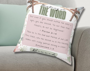 以下が含まれることがあります： 「Resting on the Word」のテキストと聖書の詩が書かれた正方形の装飾枕。枕はピンクとグリーンの配色で花柄のデザインが特徴です。枕は灰色のソファの上に置かれています。