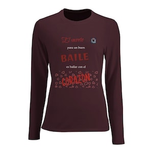 May include: Burgundy long-sleeve shirt with the Spanish text "El secreto para un buen baile es bailar con el corazón" in red. The shirt features a small square design near the shoulder and heart graphics.