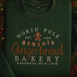 May include: A dark green sweatshirt with the embroidered text "North Pole Gingerbread Bakery" in white and brown. The design includes gingerbread men, houses, and stars. The phrase "Handmade with Love" is also embroidered.