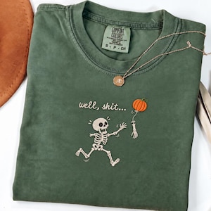 May include: An olive green short-sleeve t-shirt featuring an embroidered graphic of a skeleton reaching for a floating orange pumpkin. The text "well, shit..." is embroidered above the skeleton. The shirt has a Comfort Colors brand label.