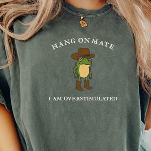 May include: Dark green t-shirt with the text "HANG ON MATE" above a cartoon frog wearing a cowboy hat and boots. The text "I AM OVERSTIMULATED" is below the frog. The shirt is a relaxed fit.