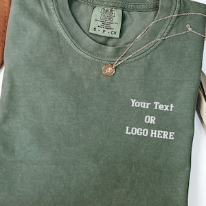 May include: A sage green short-sleeve t-shirt with a crew neckline and a Comfort Colors brand label at the inner collar. The front features the white printed text Your Text OR LOGO HERE. A gold-toned pendant necklace rests on the fabric.