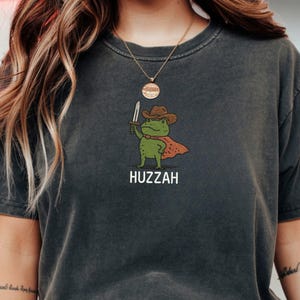May include: Dark gray t-shirt featuring a cartoon frog wearing a cowboy hat and cape, holding a sword. The word "Huzzah" is printed below the frog illustration. The shirt has a vintage, worn look.
