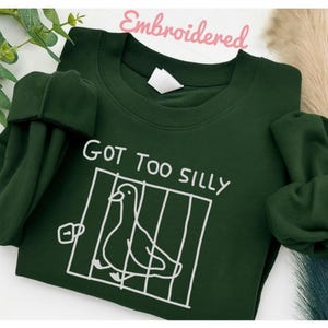 以下が含まれることがあります： 「GOT TOO SILLY」の文字と、鳥かごの中にいる鳥の白いグラフィックが描かれた、濃い緑色のスウェットシャツ。「Embroidered」の文字がピンク色で書かれています。スウェットシャツは長袖で、丸い襟ぐりです。