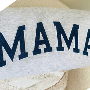 May include: A light grey heathered sweatshirt featuring the word MAMA in large, dark navy blue block letters. The letters are appliqued onto the fabric with a visible stitched border, creating a textured and bold appearance across the chest.