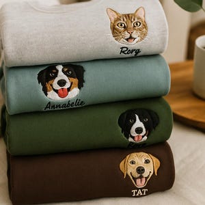 May include: A pile of folded sweatshirts in shades of grey, green, and brown, each embroidered with a pet's face and name. The top sweatshirt features a cat named "Rory". The others have dog faces and names.