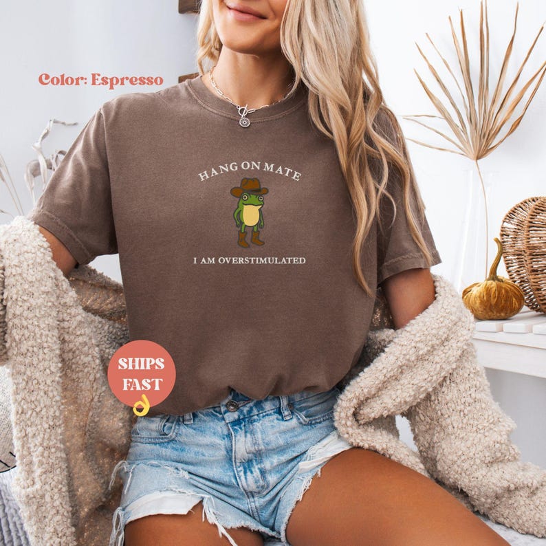May include: Espresso-colored t-shirt with the text "HANG ON MATE I AM OVERSTIMULATED" and a cartoon frog wearing a cowboy hat and boots. The shirt is a short-sleeved, crew neck style.