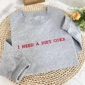 May include: A gray sweatshirt with the text "I NEED A DIET COKE" embroidered in red.