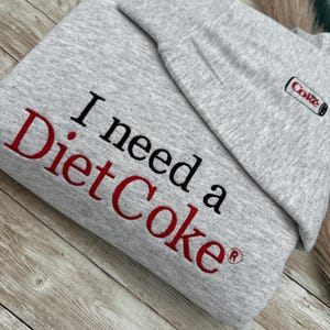 May include: Gray sweatshirt with the text "I need a Diet Coke" in red and black lettering. A small red and white "Coke" logo is on the right side of the sweatshirt.