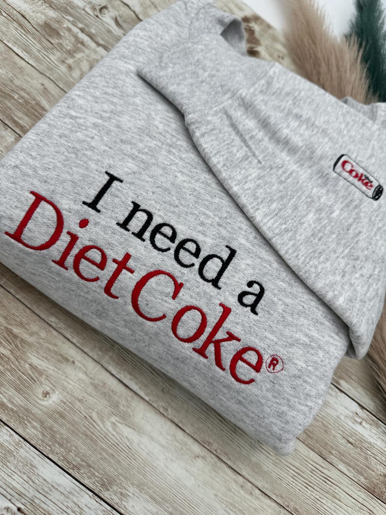May include: Gray sweatshirt with the text "I need a Diet Coke" in red and black lettering. A small red and white "Coke" logo is on the right side of the sweatshirt.