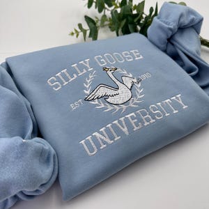 May include: A light blue sweatshirt with white embroidered text that reads "Silly Goose University Est. 1910" and features a white embroidered goose with a crown.