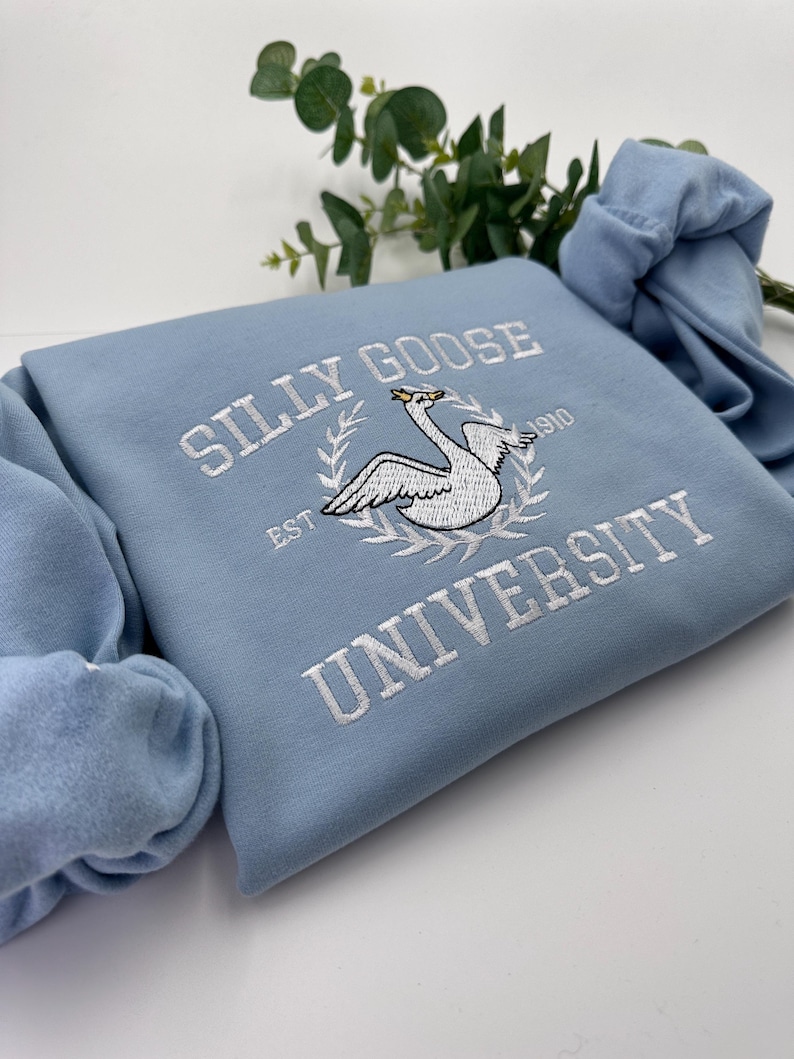 May include: A light blue sweatshirt with white embroidered text that reads "Silly Goose University Est. 1910" and features a white embroidered goose with a crown.