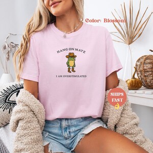 May include: A light pink t-shirt with the text "HANG ON MATE" above a cartoon frog wearing a cowboy hat and boots. The text "I AM OVERSTIMULATED" is below the frog. The shirt is a blossom color.