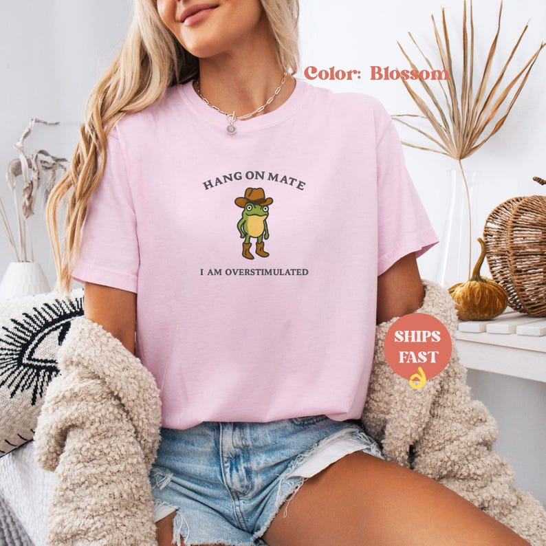 May include: A light pink t-shirt with the text "HANG ON MATE" above a cartoon frog wearing a cowboy hat and boots. The text "I AM OVERSTIMULATED" is below the frog. The shirt is a blossom color.