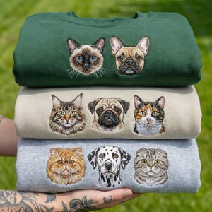 May include: Three sweatshirts in green, beige, and grey, stacked and embroidered with pet portraits. The top one features a Siamese cat and a French bulldog. The middle one has a tabby cat, a pug, and a calico cat. The bottom one has a Persian cat, a Dalmatian, and a Scottish Fold cat.