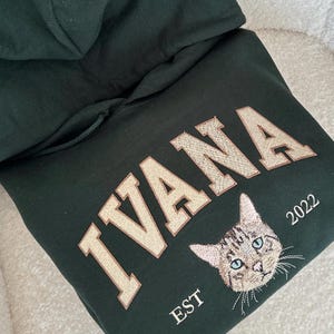 May include: A dark green hoodie with the word "IVANA" embroidered in gold on the front. Below the word is a small embroidered image of a cat and the text "EST 2022".
