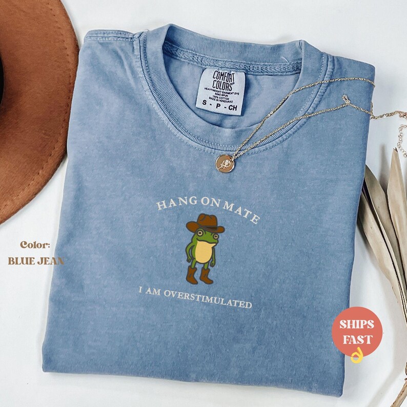 May include: Blue jean Comfort Colors t-shirt with a graphic of a frog wearing a cowboy hat and boots. Text reads "HANG ON MATE" and "I AM OVERSTIMULATED". A gold necklace is draped across the shirt.