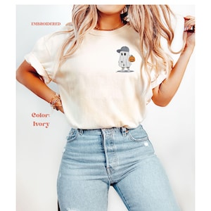 May include: Ivory-colored t-shirt with an embroidered ghost wearing a baseball cap and holding a pumpkin. The shirt is paired with light blue jeans. Text on the shirt reads "EMBROIDERED" and "Color: Ivory".