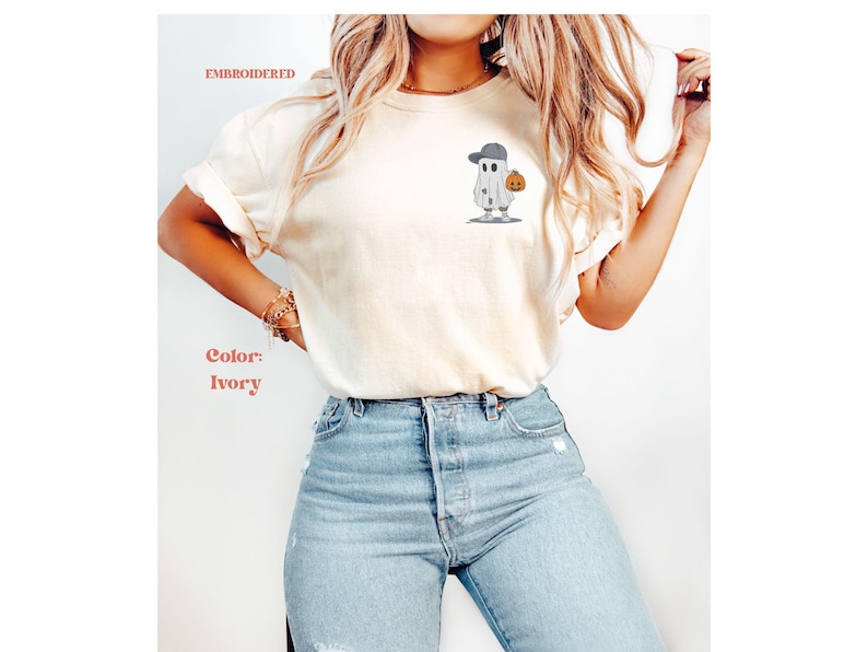 May include: Ivory-colored t-shirt with an embroidered ghost wearing a baseball cap and holding a pumpkin. The shirt is paired with light blue jeans. Text on the shirt reads "EMBROIDERED" and "Color: Ivory".