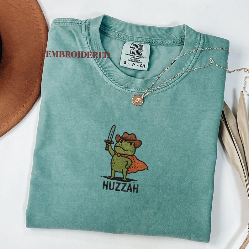 May include: A teal Comfort Colors t-shirt with an embroidered design of a green frog wearing a cowboy hat and cape, holding a sword. The word "Huzzah" is printed below the frog. A silver necklace with a pendant is draped across the shirt.