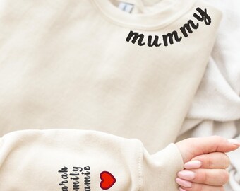 May include: A cream colored sweatshirt with the word "Mummy" embroidered in black on the neckline. The left sleeve has the names "Sarah, Emily, Jamie" embroidered in black with a red heart.