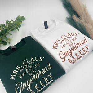 May include: Two sweatshirts, one white and one dark green, with the text "MRS. CLAUS Gingerbread BAKERY" and a gingerbread man graphic. The text includes "THE NORTH POLE EST 1898" and "REAL GOOD COOKIES".