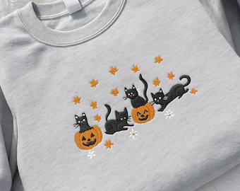 Puede incluir: Una sudadera gris claro con un diseño bordado de Halloween. El diseño incluye cuatro gatos negros, cada uno con una pose diferente, con tres gatos sentados en calabazas naranjas talladas. Estrellas naranjas y flores blancas completan el diseño.