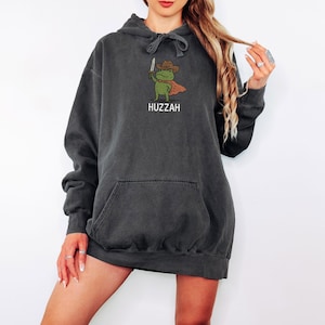 May include: Dark gray hoodie with a front pocket and drawstring hood. The front features a cartoon frog wearing a cowboy hat and cape, holding a sword. The word "Huzzah" is printed below the frog in white.