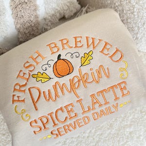 Pode incluir: Almofada retangular creme com texto bordado: "FRESH BREWED Pumpkin SPICE LATTE SERVED DAILY." O design inclui uma ab&oacute;bora laranja e folhas amarelas. A almofada est&aacute; sobre uma superf&iacute;cie branca texturizada.
